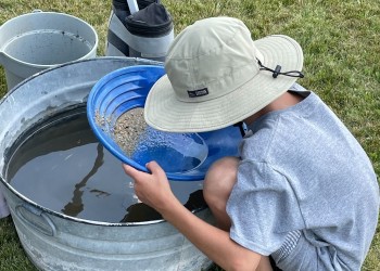A child in a gray shirt squats before a water-filled galvanized bin holding a blue plastic gold.pan. 