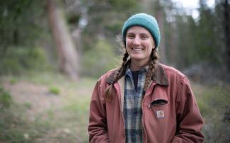 Woman in green stocking cap, coat and flannel stands in front of trees